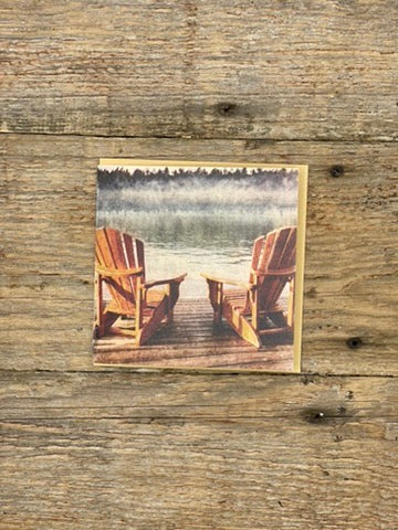 Wooden postcard with two Adirondack chairs on a dock over a lake, placed on a wooden surface.