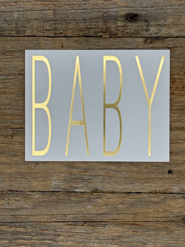 Card with gold 'BABY' text on a gray background, placed on wooden surface