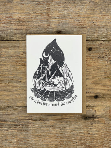 Card with black and white illustration of a campfire scene on a wooden surface