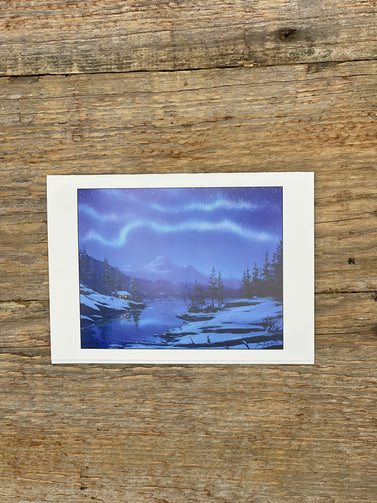 Card with painting of a snowy landscape with mountains and a lake on a wooden surface.