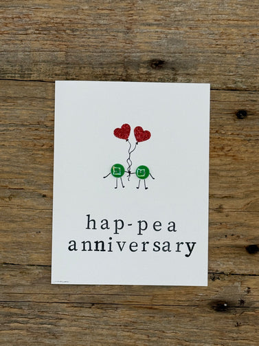 Card with two green peas holding red heart balloons and 'hap-pea anniversary' text on a wooden surface.
