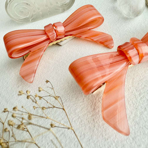 Pair of coral bow-shaped hair clips on a light background with dried flowers.