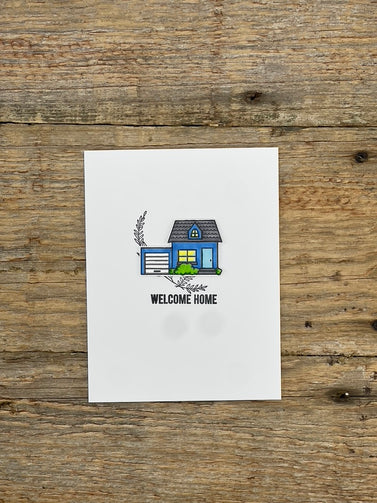 White card with a blue house illustration and 'Welcome Home' text on a wooden surface