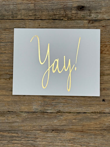 Card with 'Yay!' in gold on a gray background, placed on wooden surface