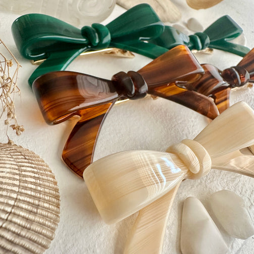 Three hair bows in green, brown, and beige on a textured white surface with shells.
