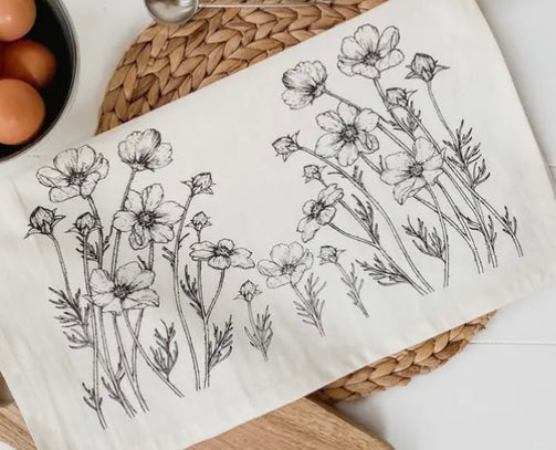 A beige organic cotton tea towel with a black floral print, displayed on a white surface surrounded by green foliage.
