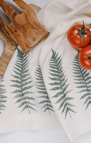 A beige organic cotton tea towel with a black floral print, displayed on a white surface surrounded by green foliage.