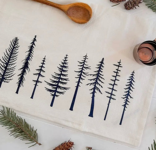 A beige organic cotton tea towel with a black floral print, displayed on a white surface surrounded by green foliage.