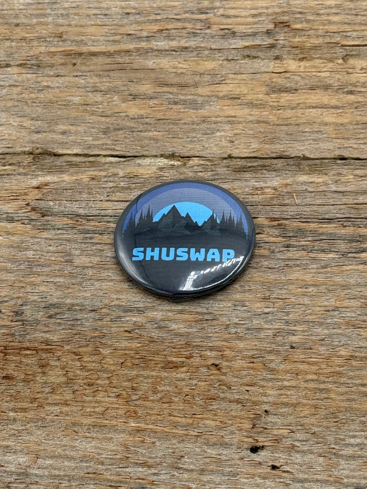 Button badge with 'SHUSWAP' text and mountain design on a wooden surface