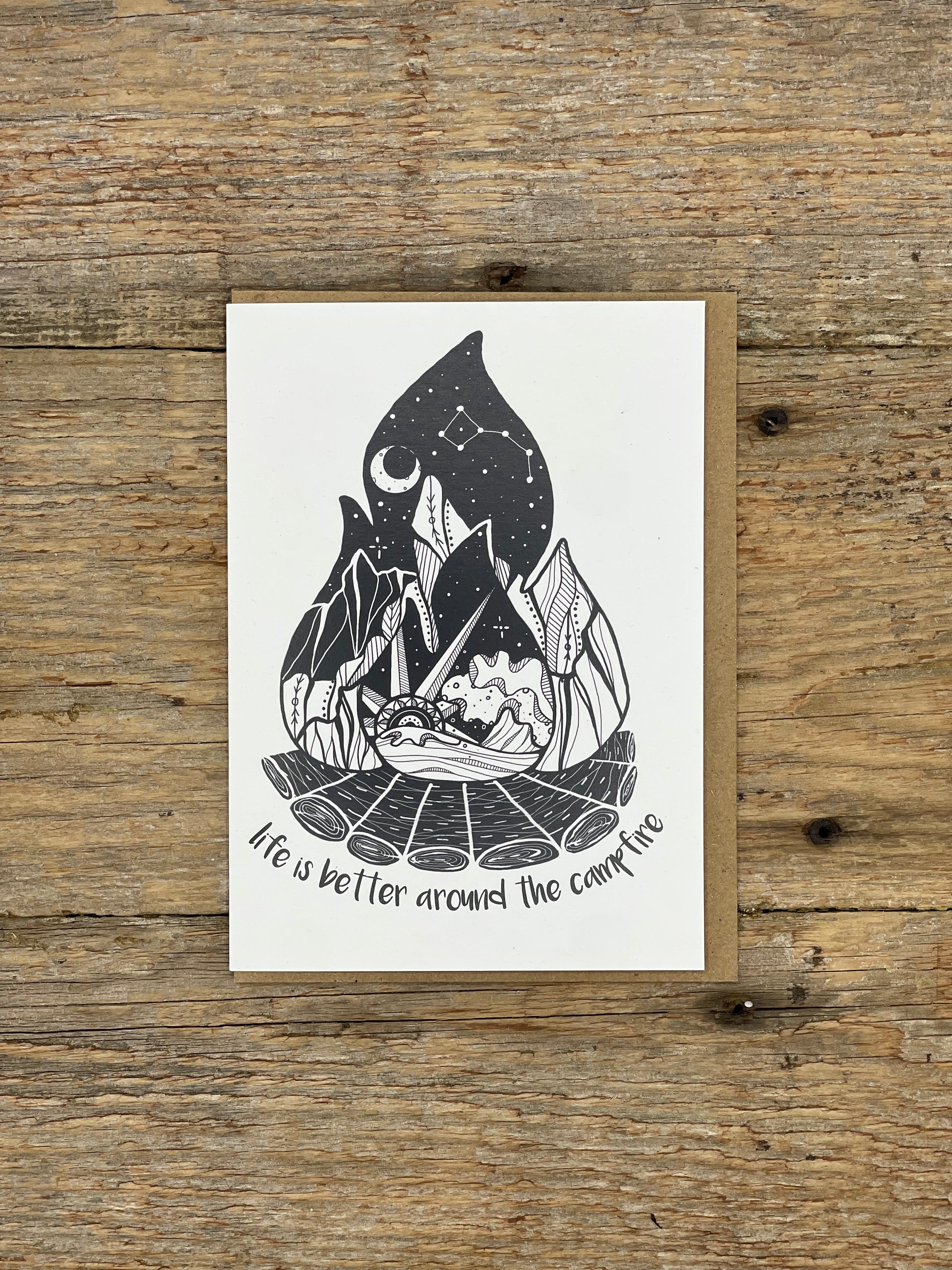 Card with black and white illustration of a campfire scene on a wooden surface
