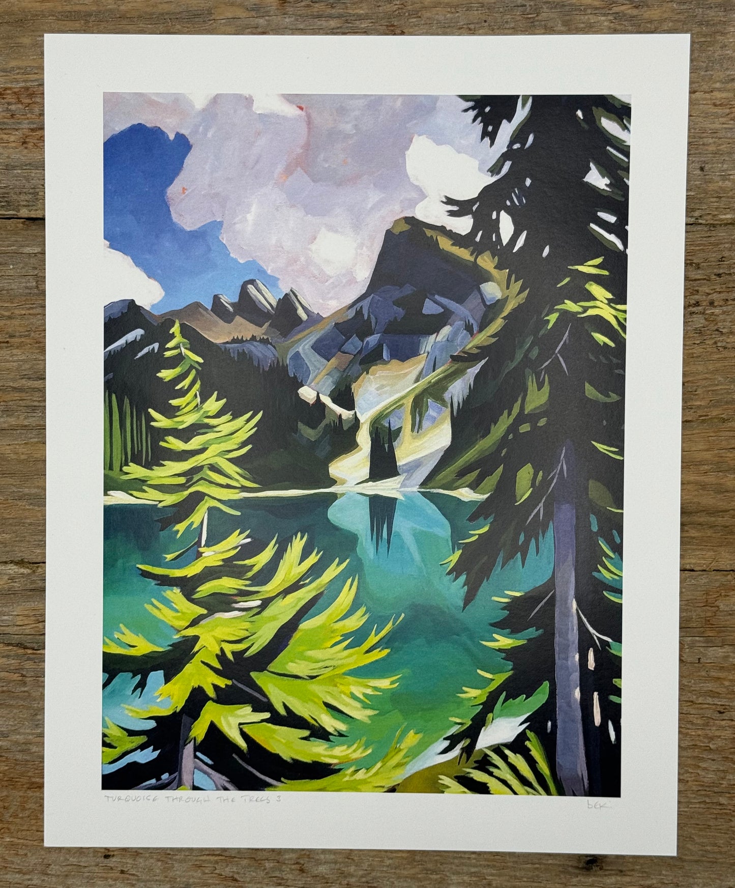 Framed artwork of a mountain landscape with trees and water on a wooden surface