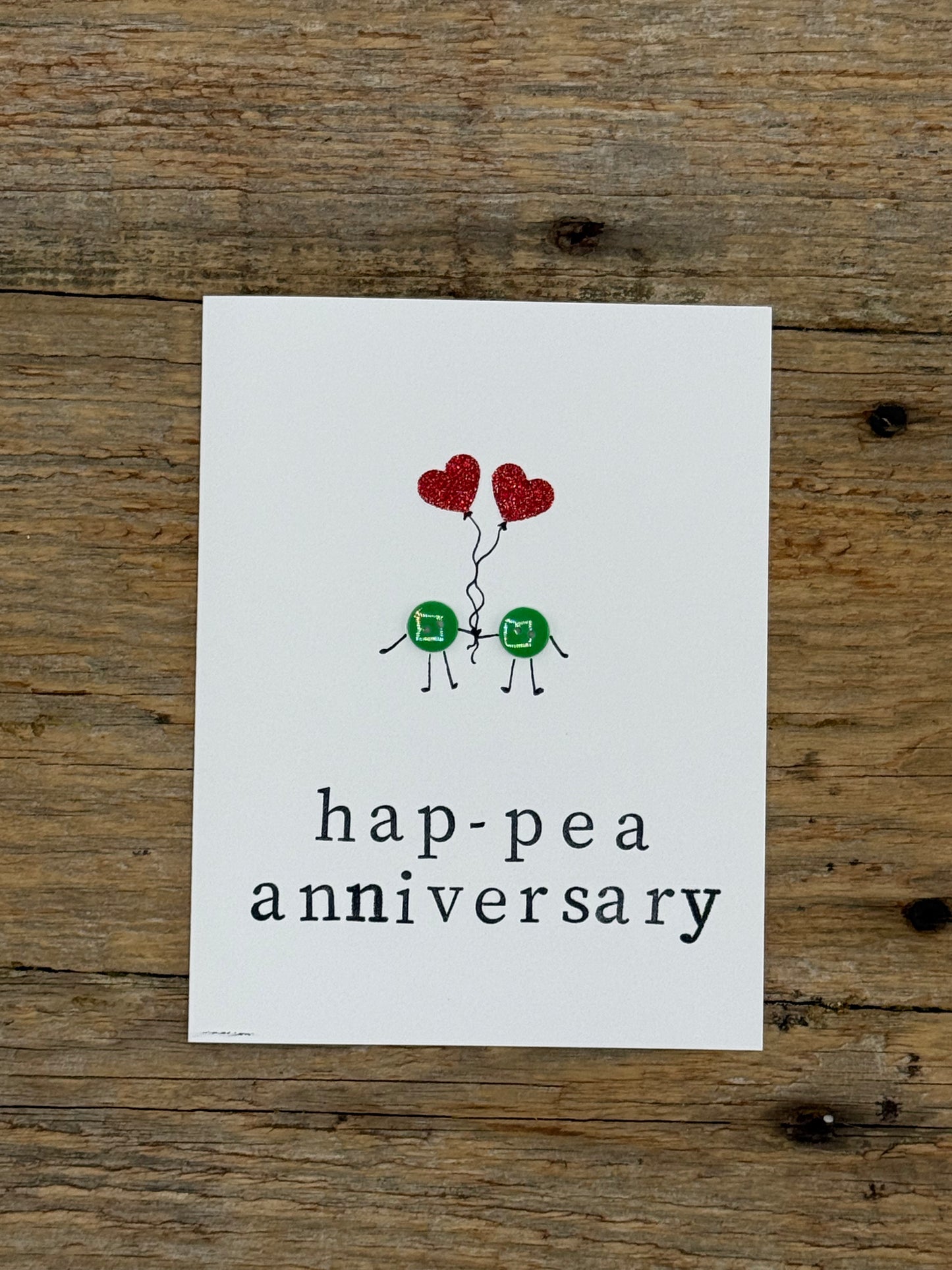 Card with two green peas holding red heart balloons and 'hap-pea anniversary' text on a wooden surface.