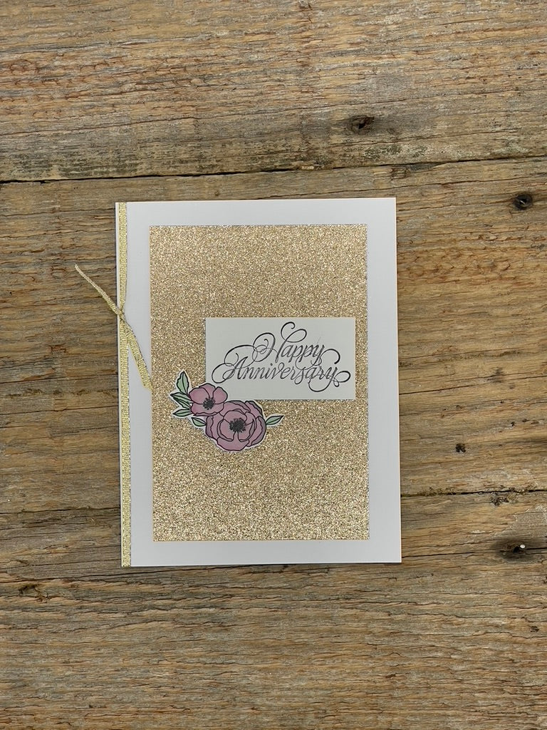 Greeting card with 'Happy Anniversary' text and floral design on a glittery background, placed on a wooden surface.