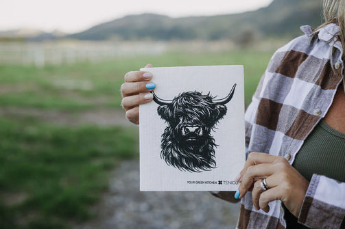 Person holding a card with a black and white illustration of a Highland cow in a field.