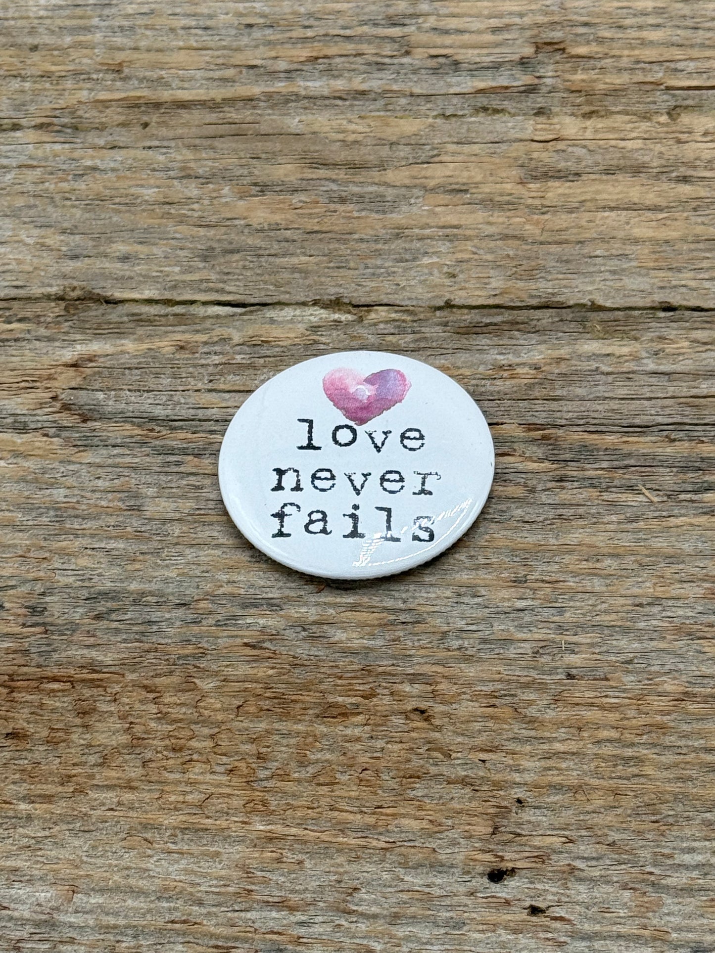 magnet with 'love never fails' text and pink heart on wooden surface