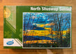 Jigsaw puzzle box titled 'North Shuswap Sunset' by Venus Puzzle on a wooden surface.