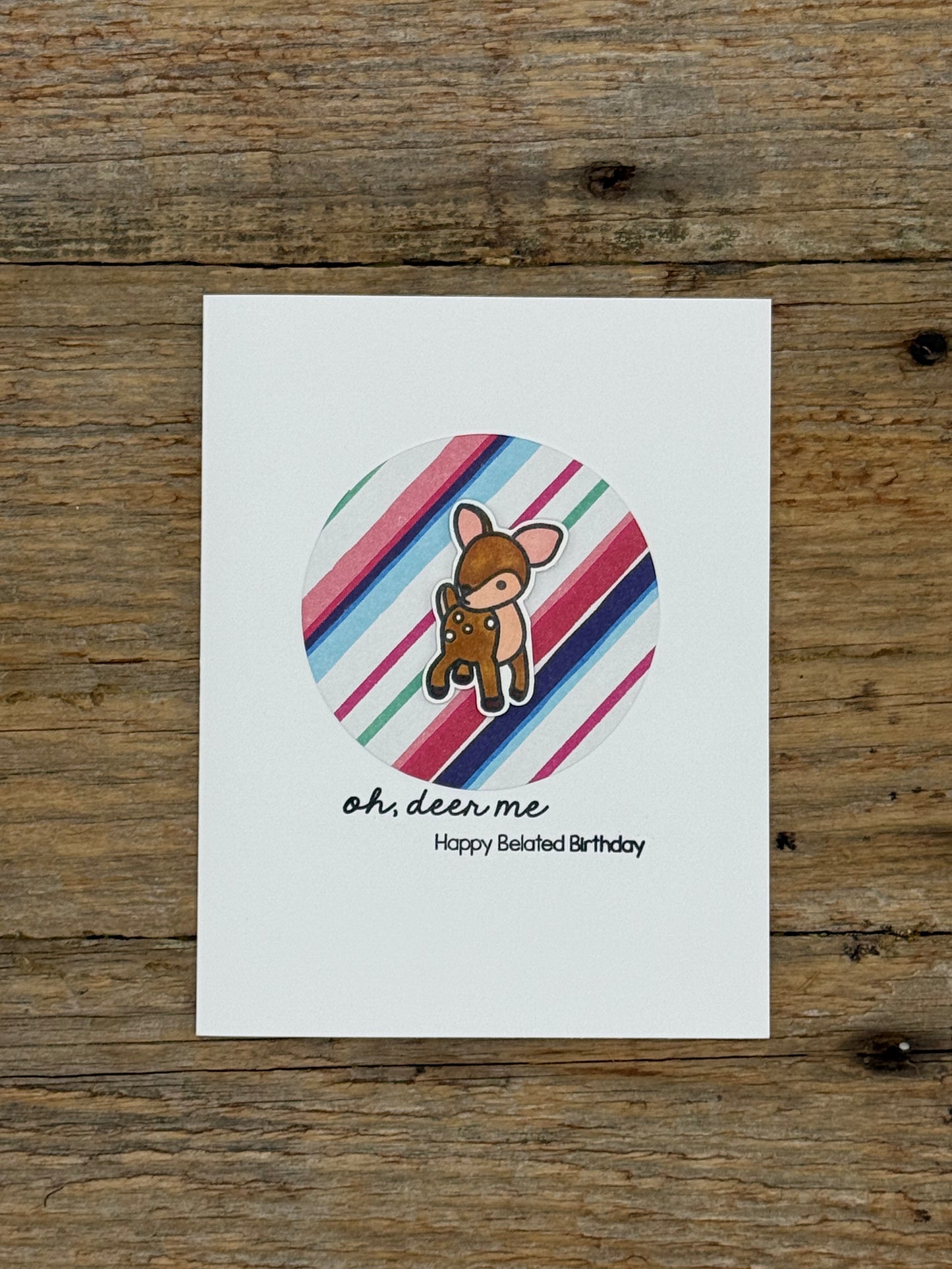 Card with a cartoon deer on a striped background, placed on a wooden surface.