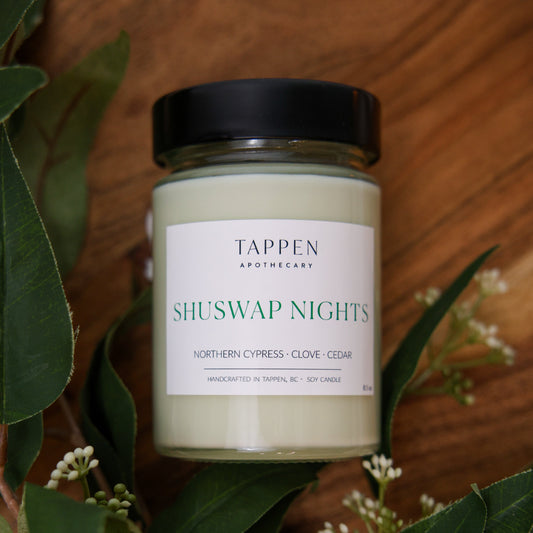 Candle jar labeled 'Shuswap Nights' by Tappen Apothecary on a wooden surface with green leaves.
