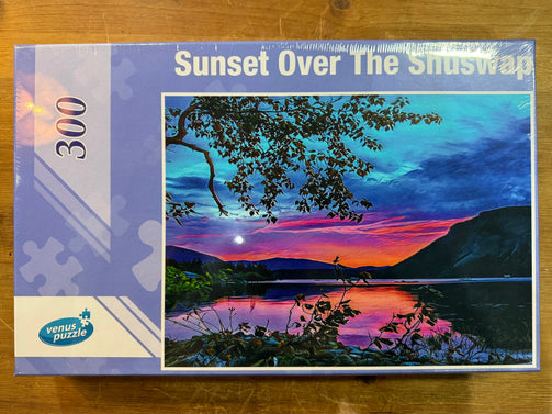 Jigsaw puzzle box titled 'Sunset Over The Shuswap' by Venus Puzzle on a wooden surface.