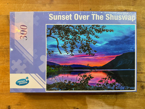 Jigsaw puzzle box titled 'Sunset Over The Shuswap' with a scenic illustration on a wooden surface.