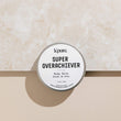 A metal tin container with a white label that reads 'SUPER OVERACHIEVER Body Balm' on a marble background.