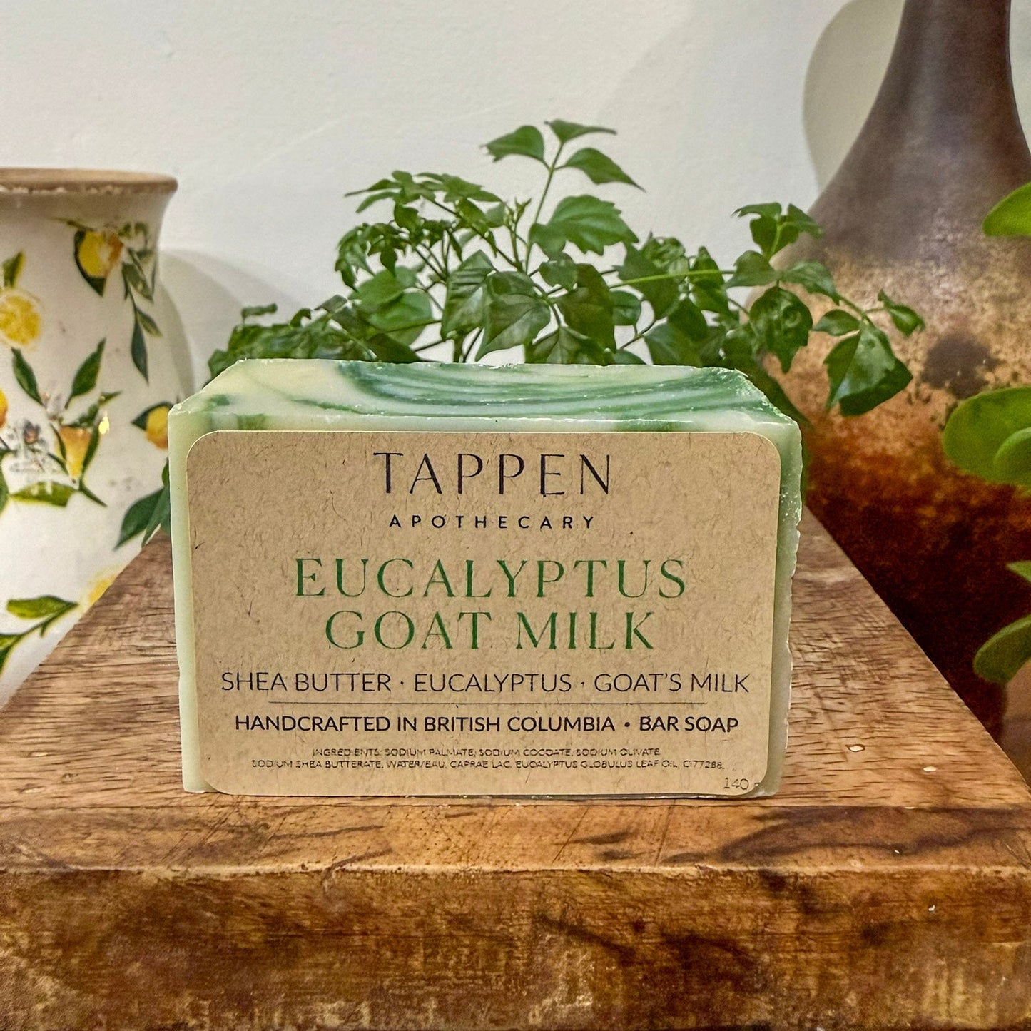 Bar of Tappen Apothecary Eucalyptus Goat Milk soap on a wooden surface with decorative vases and plants in the background.