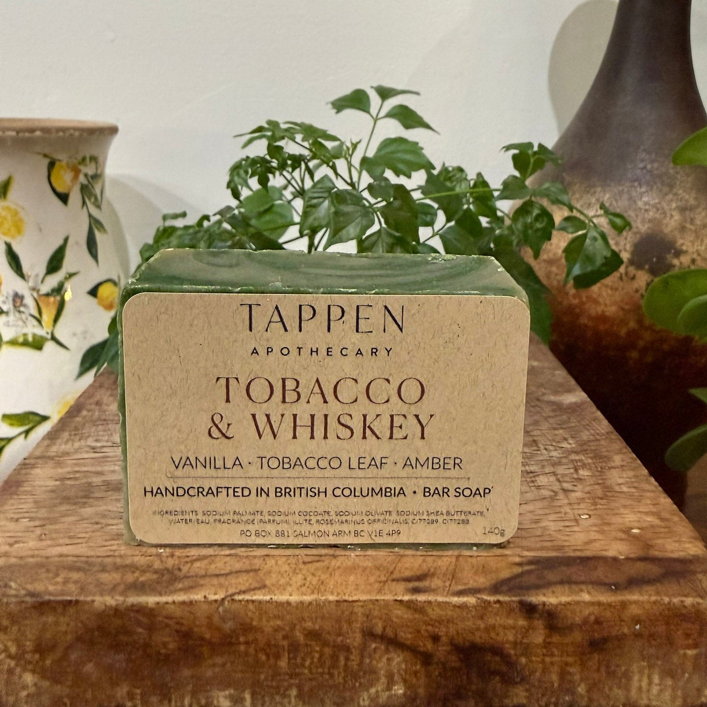 Bar of Tappen Apothecary Tobacco & Whiskey soap on a wooden surface with plants in the background.