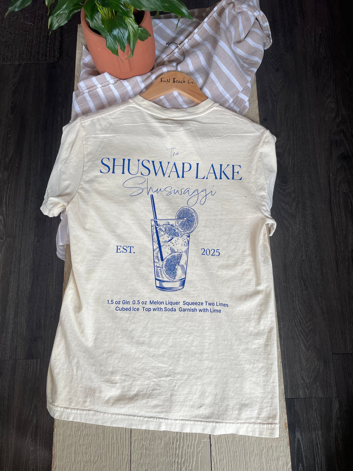 An off-white graphic t-shirt, featuring a blue print of a Shuswap Lake cocktail and text of the drink recipe.