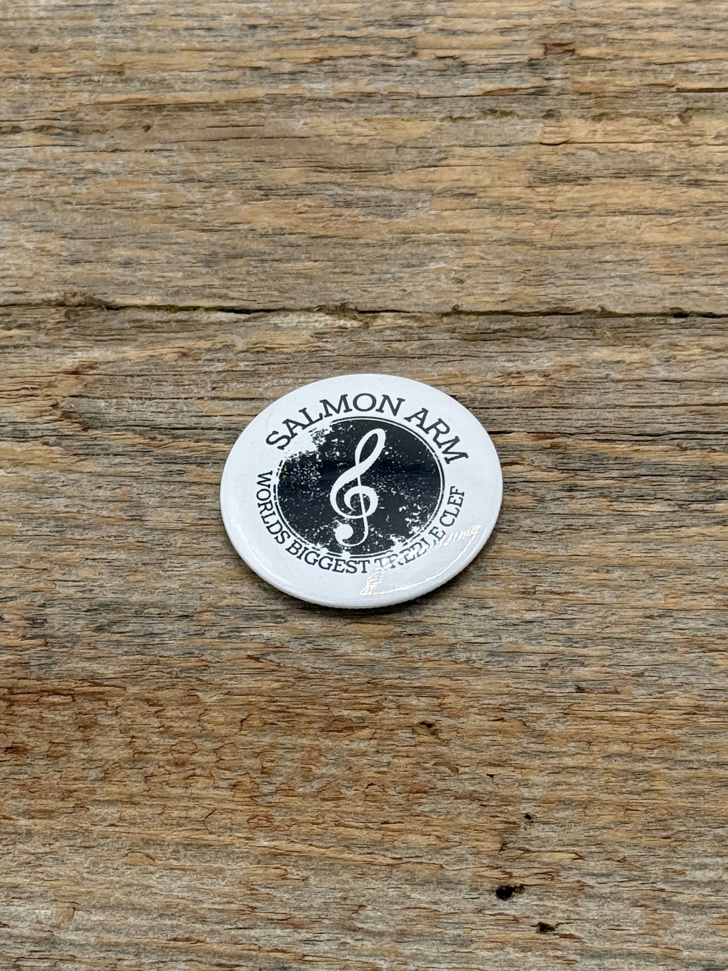 Round metal badge with 'Salmon Arm World's Biggest Treble clef' text and musical symbol on a wooden surface
