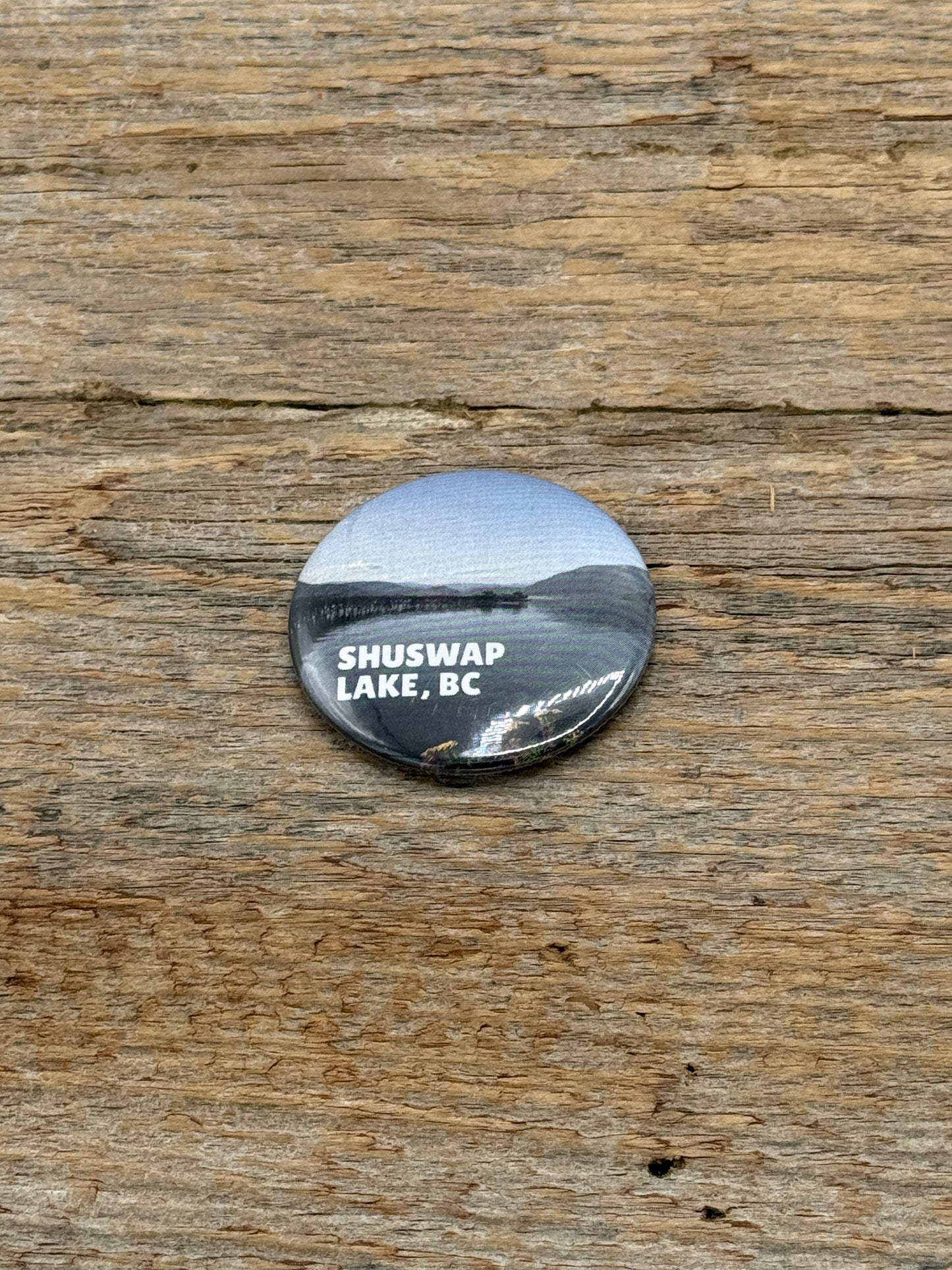Round metal badge with 'Shuswap Lake, BC' text on a wooden surface