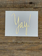 Card with 'Yay!' in gold on a gray background, placed on wooden surface