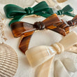 Three hair bows in green, brown, and beige on a textured white surface with shells.