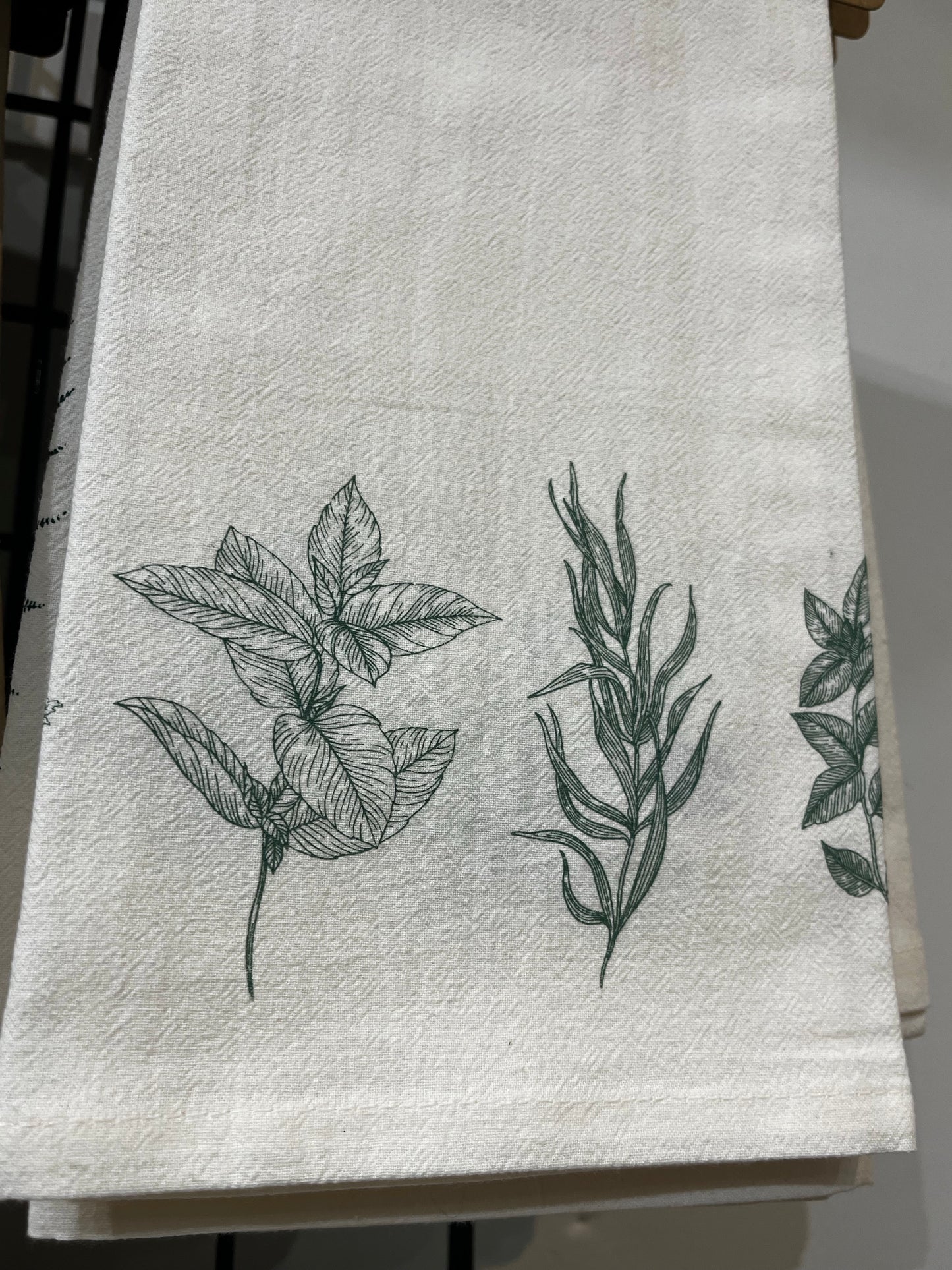 A beige organic cotton tea towel with a black floral print, displayed on a white surface surrounded by green foliage.