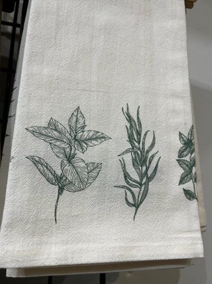 A beige organic cotton tea towel with a black floral print, displayed on a white surface surrounded by green foliage.