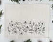 A beige organic cotton tea towel with a black floral print, displayed on a white surface surrounded by green foliage.