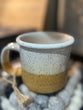 A ceramic pottery mug with a two-tone design, featuring a solid beige color at the bottom and a speckled white at the top, with a handle.